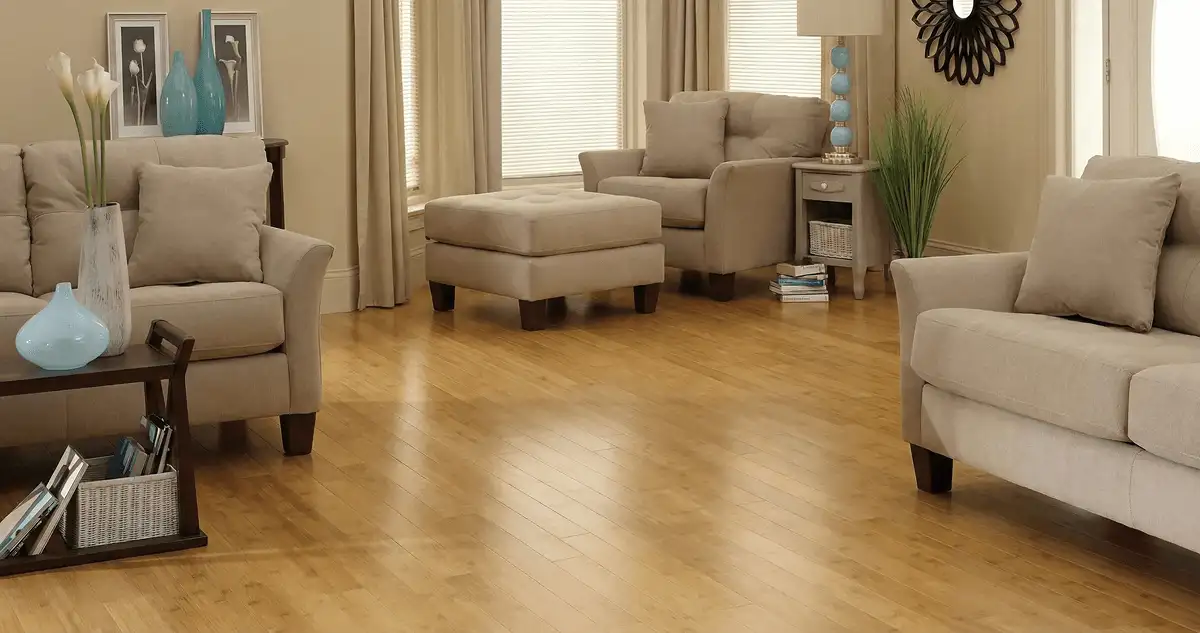A living room with professionally installed bamboo flooring, beige sofas, a matching ottoman, a side table with a blue lamp, vases, framed pictures, and large windows with blinds and curtains in Sioux Falls, SD